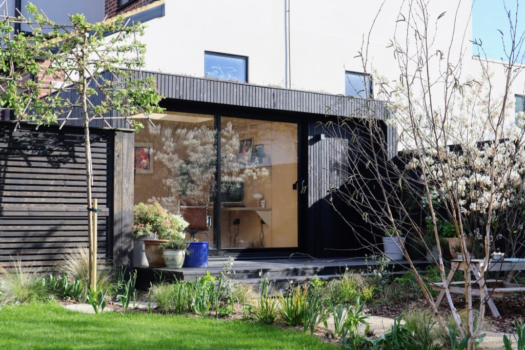 A black timber-clad garden room with sliding glass doors, set in a planted garden