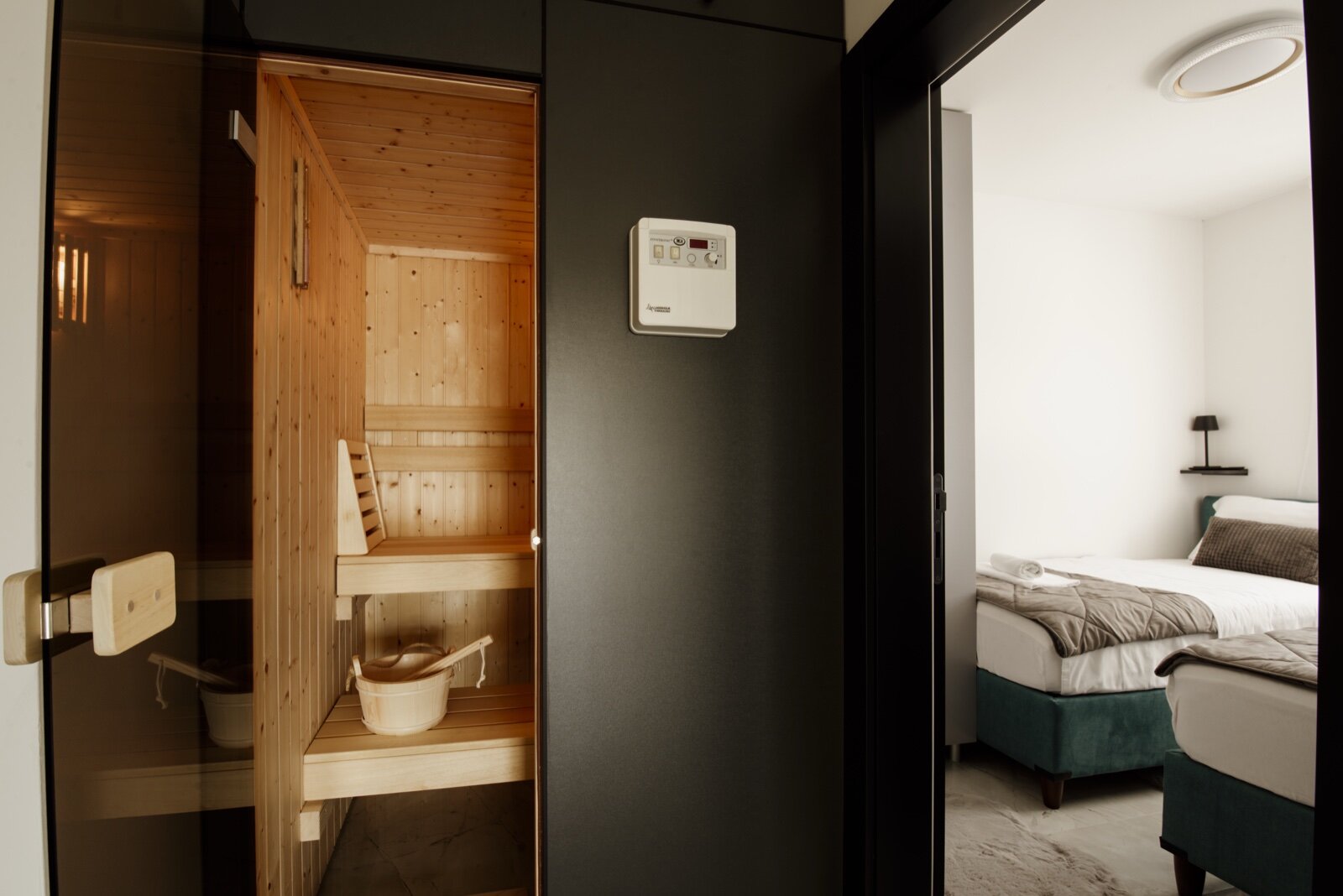 Sauna cabin with timber benches, seen through a glass door, with the control panel mounted on the wall outside