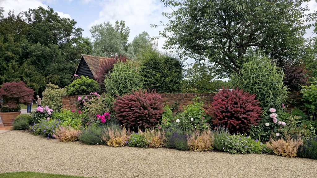 Layered shrub and perennial border beside a gravel path, with a timber garden building behind a brick wall