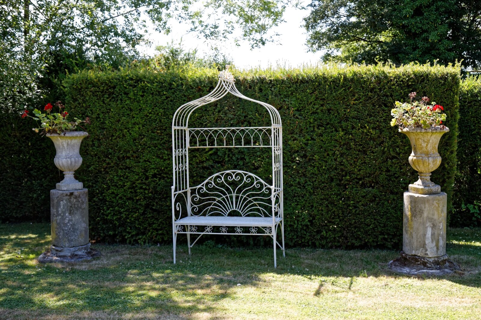 White metal bench arbour against a clipped hedge between two stone urns