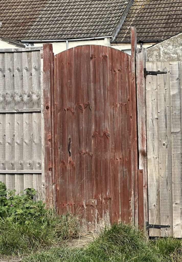 Arched wooden side gate stained red-brown, set between panel fences with black tee hinges