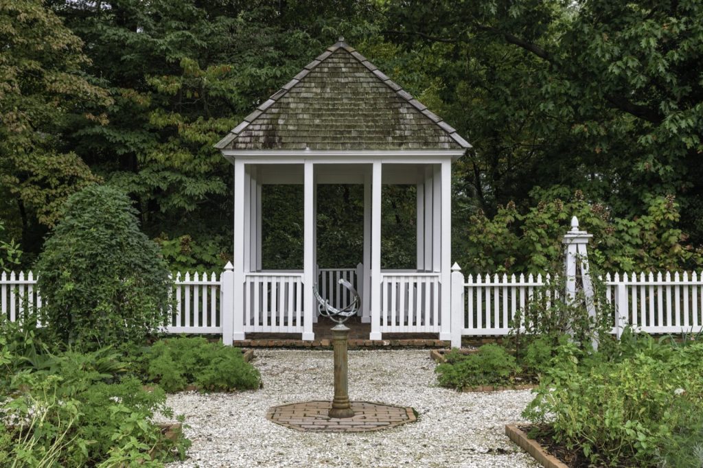 White timber garden gazebo with a shingle roof behind a picket fence