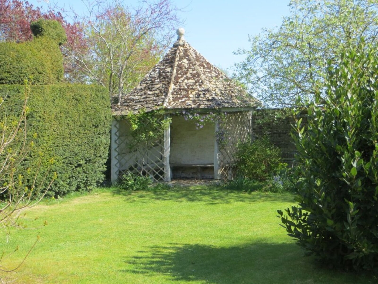 A corner garden shelter with a shingled conical roof, trellis sides and a built-in bench, set against a clipped hedge