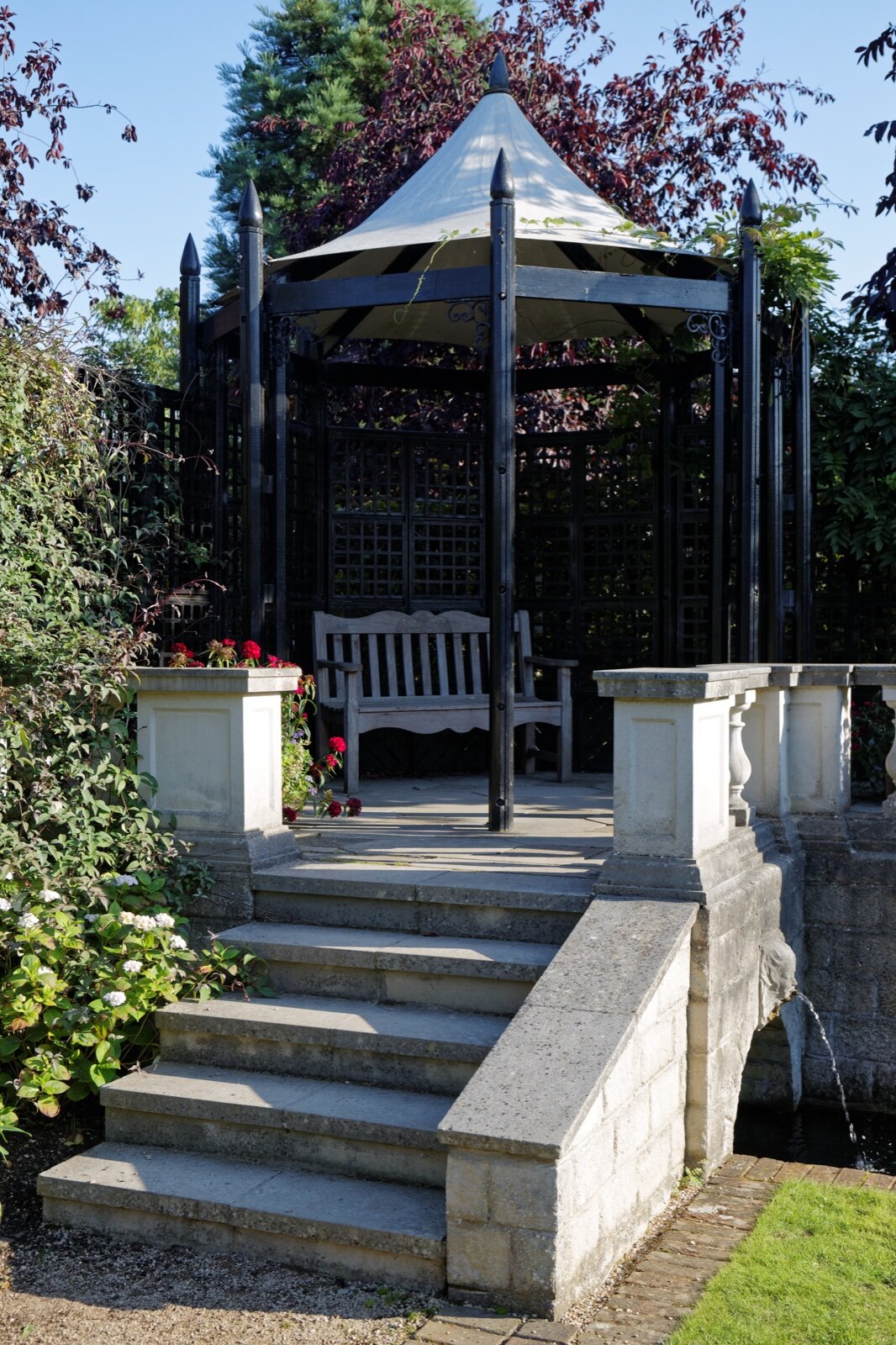A black framed garden gazebo with a fabric roof, trellis panels and a timber bench inside, reached by stone steps