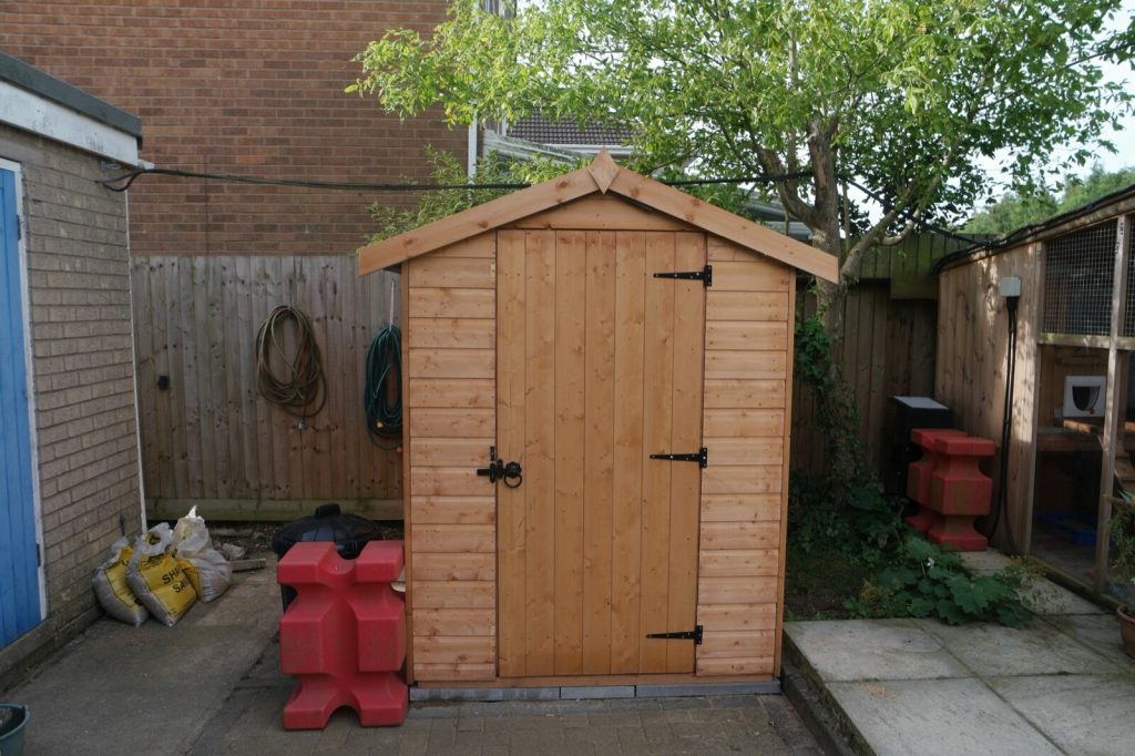 A small new apex wooden shed with a single door standing on a patio
