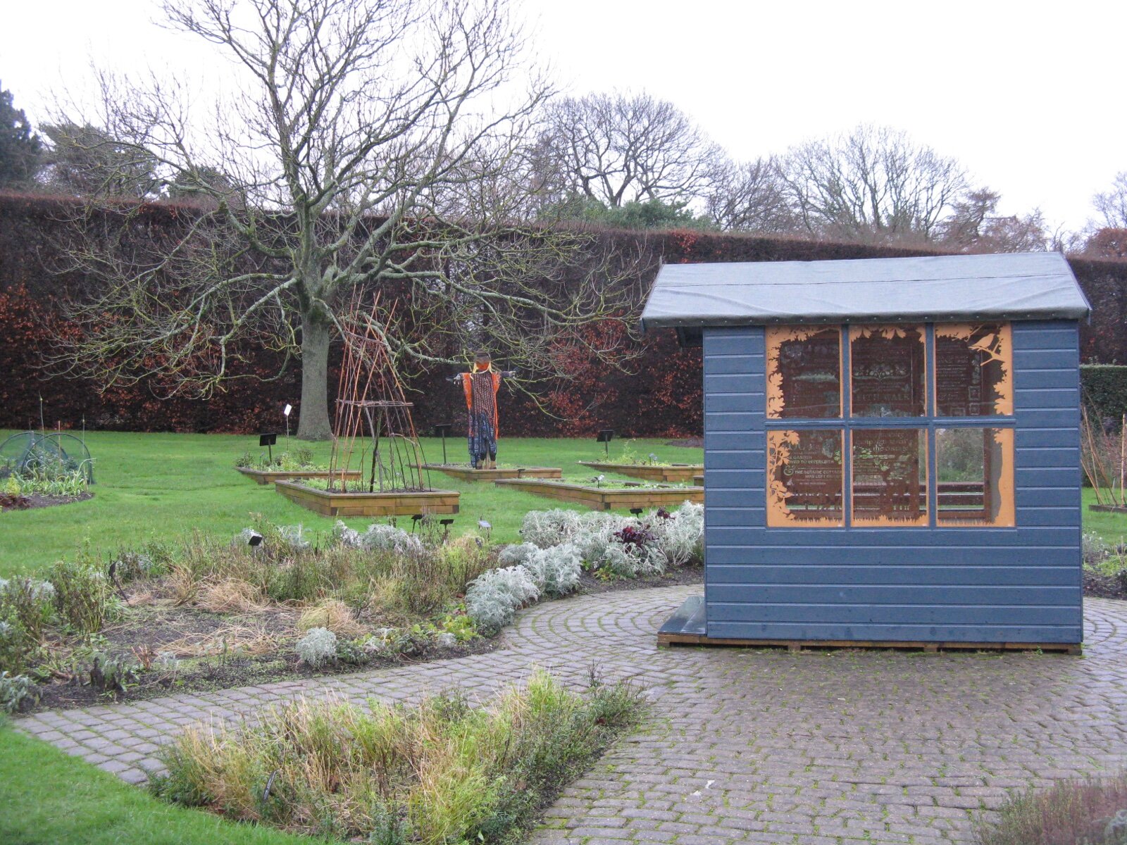 A small blue-painted pent roof shed with a wide window on a block-paved circle in a garden