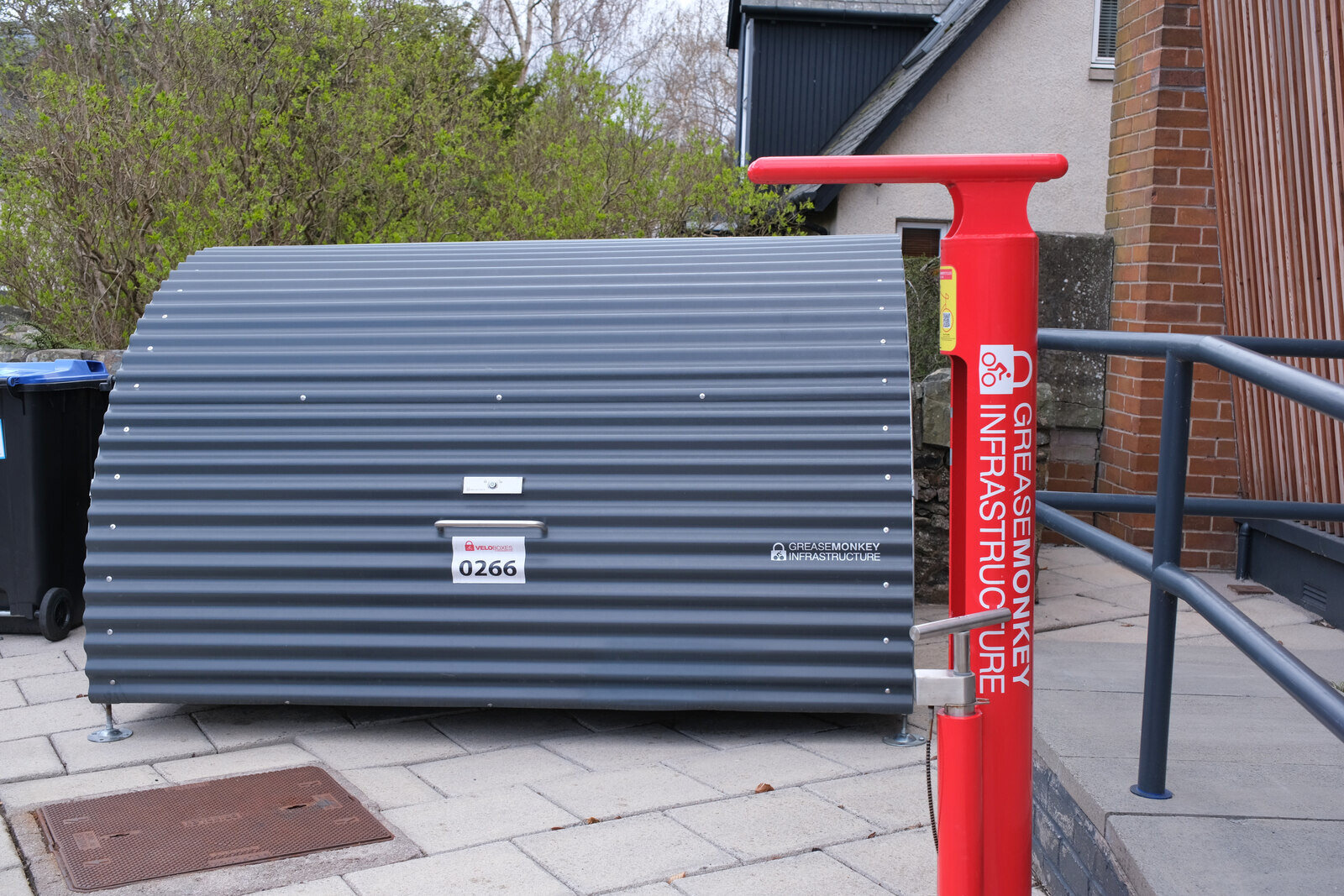 A curved grey metal bike locker on paving beside a red public bike pump