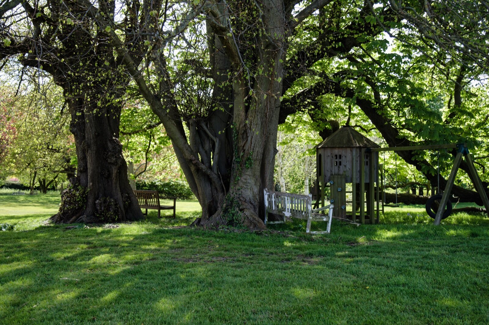 Raised wooden playhouse with a swing frame under large trees on a lawn