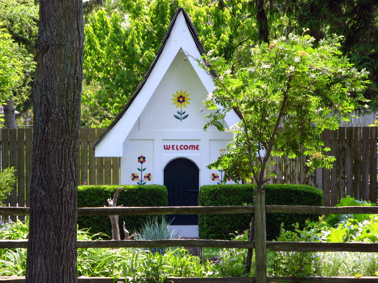 White painted children's playhouse with a steep roof behind a hedge and fence
