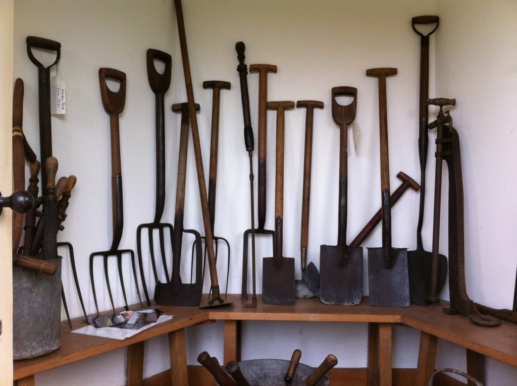 Spades, forks and rakes stored upright against a wall above a wooden bench